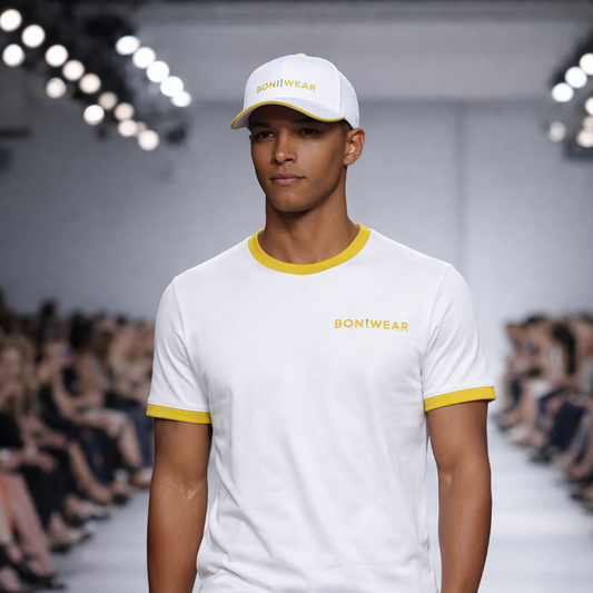 Model wearing a white t-shirt with yellow accents and a cap, walking on a fashion runway.