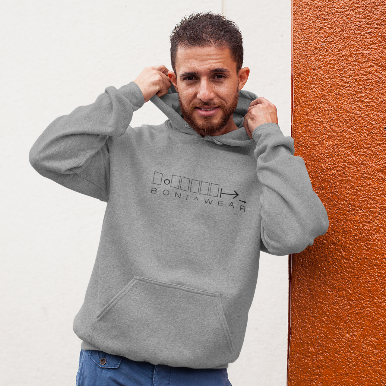 Man wearing a gray hoodie with branding against a textured wall.