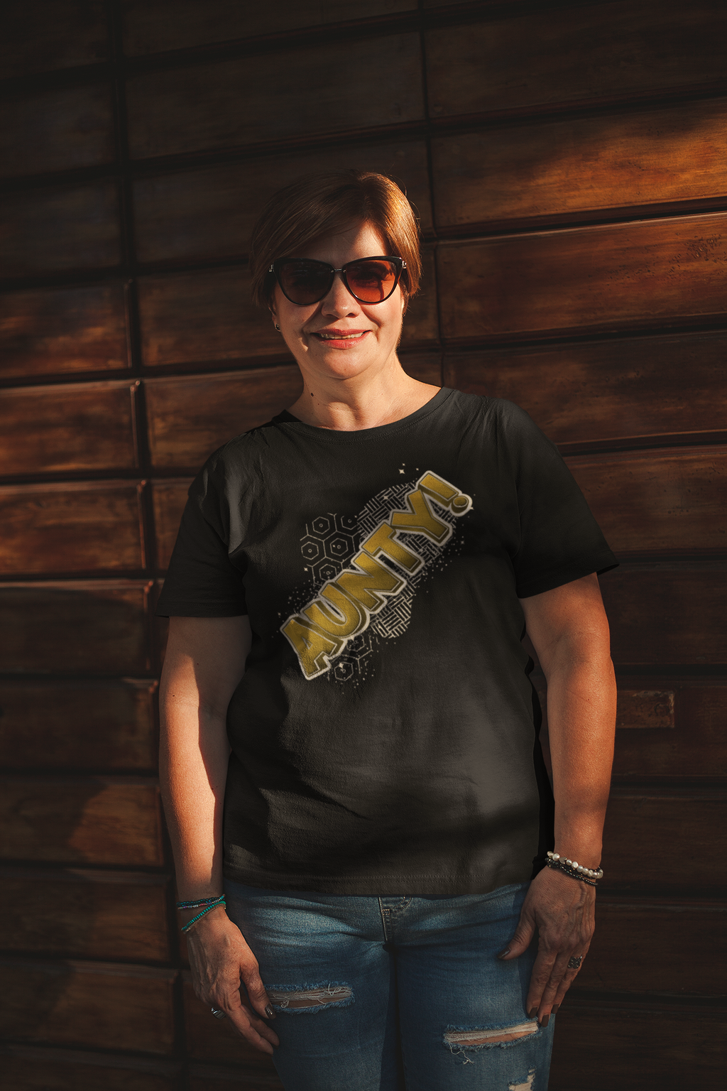 Person wearing a black t-shirt with "AUNTY" yellow text and sunglasses, standing against a wooden wall.